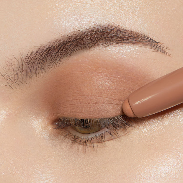 ColourPop Shadow Stix in Cocktail Party gliding across a closed eyelid, laying down a smooth matte warm nude shade along the lash line and lid, with natural brow and lashes visible to showcase creamy, blendable eyeshadow application.