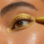 ColourPop Flex Shadow Stix applies a rich metallic gold shade on a brown eye, tracing the upper and lower lash lines and covering the lid for a bold, shimmery finish in a tight beauty close-up.